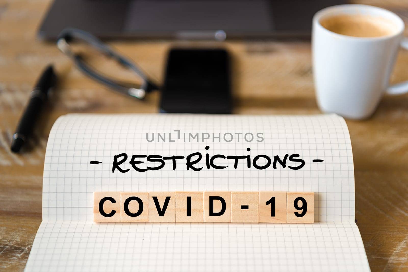 Closeup on notebook over wood table background, focus on wooden blocks with letters making Covid-19 restrictions writing. Business concept with laptop and mobile phone in a defocused background