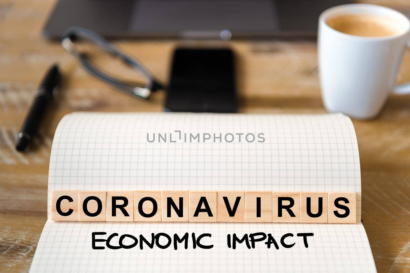Closeup on notebook over wood table background, focus on wooden blocks with letters making Coronavirus Economic Impact writing. Business concept with laptop and mobile phone in a defocused background