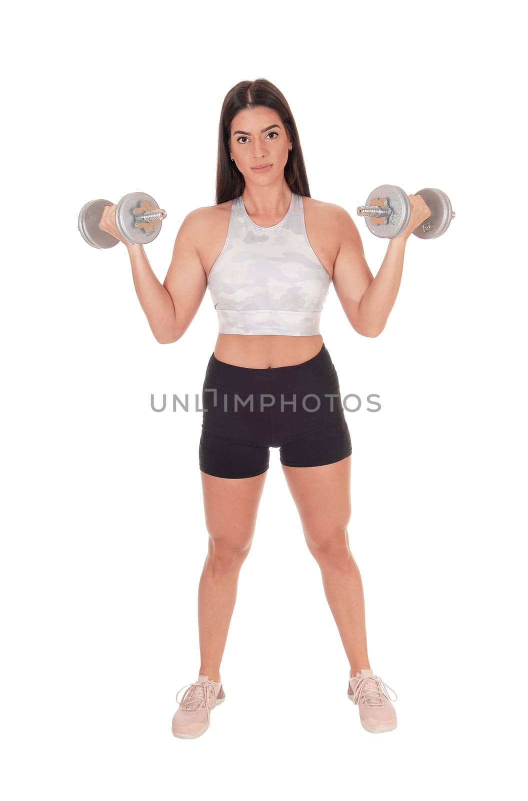A beautiful young woman standing in shorts and sports bra from the.front lifting her two dumbbells, isolated for white background.