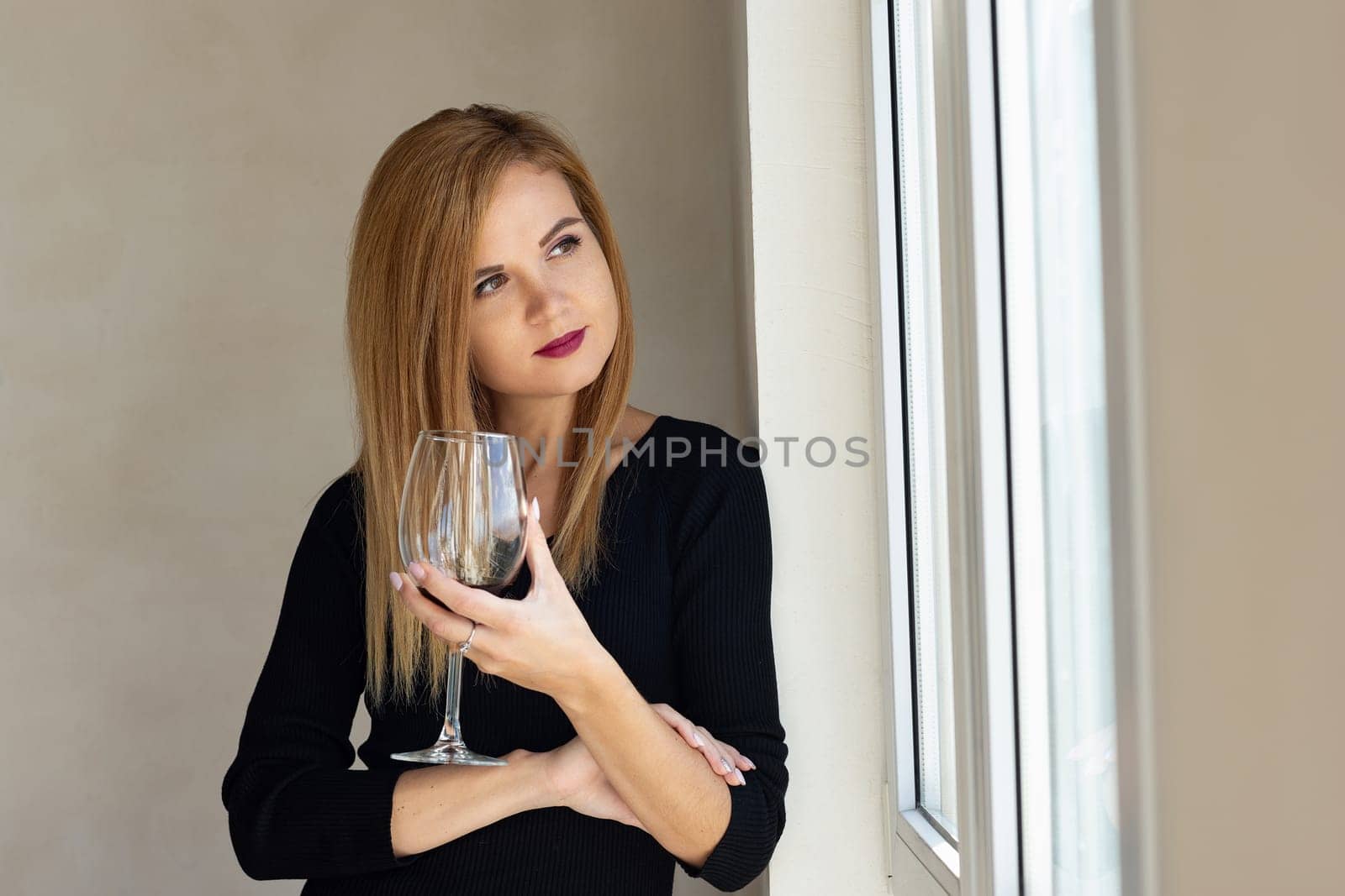 The girl stands thoughtfully at the window with a glass of red wine
