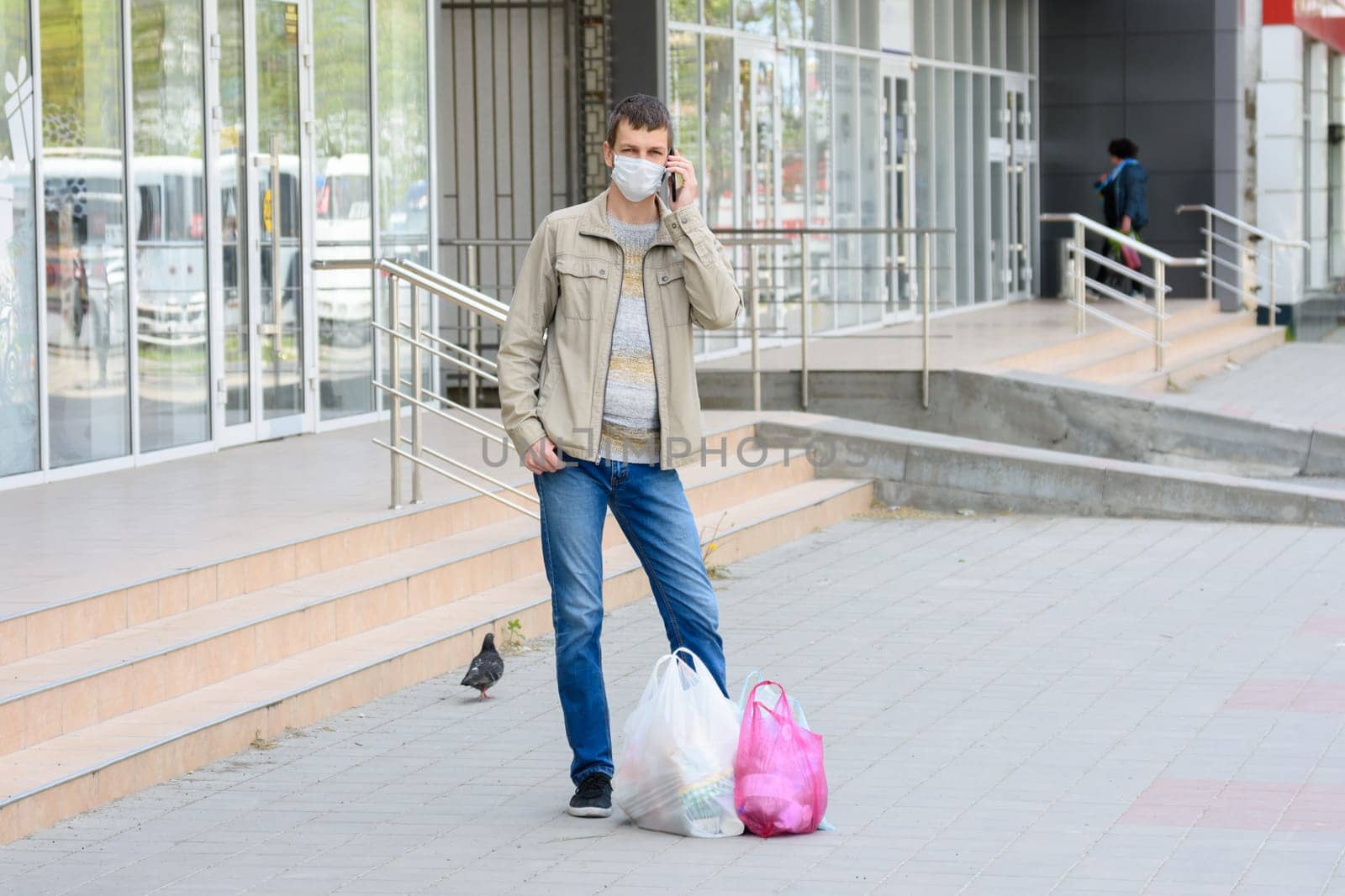 a man in a medical mask stands on the street shopping and talks on his mobile phone