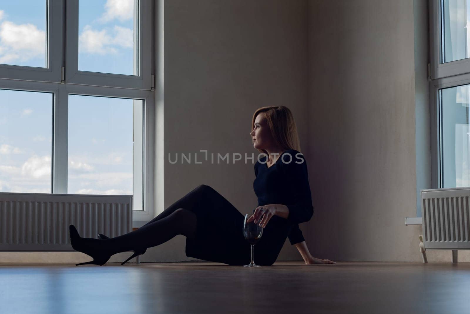 girl in evening dress and shoes sits on the floor near a large window with wine