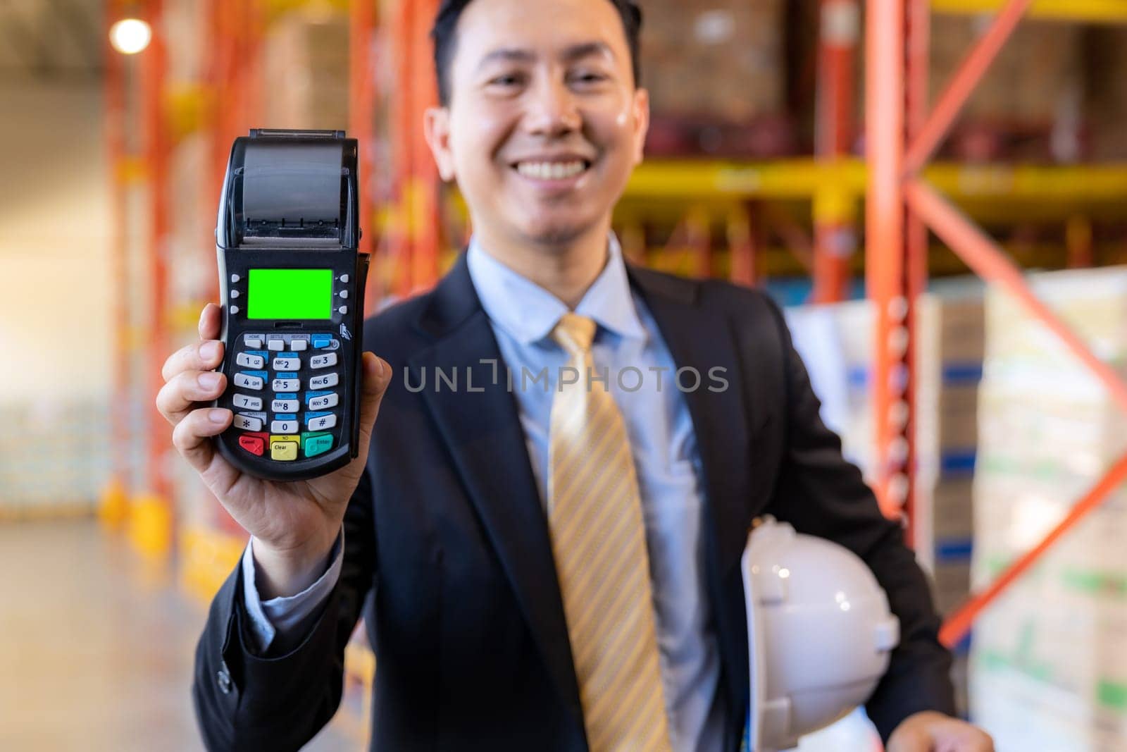 Portrait of businessman hold credit card reader in large factory and distribution warehouse environment. Business deal merger acquisitions and takeover.