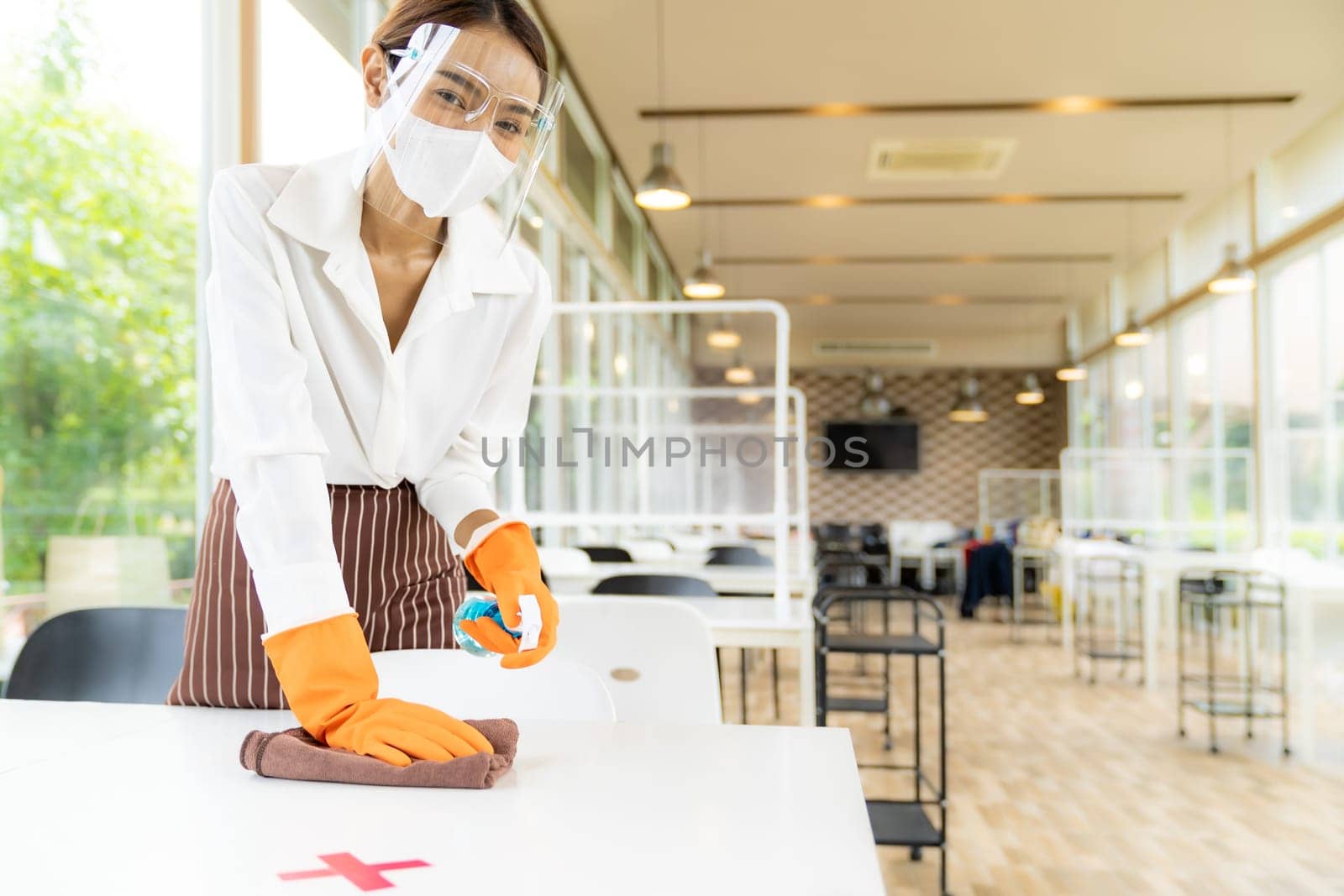 Portrait attractive asian waitress wear face mask and face shield cleaning table with alcohol and wet wipe before welcome customer. New normal hygiene restaurant concept.