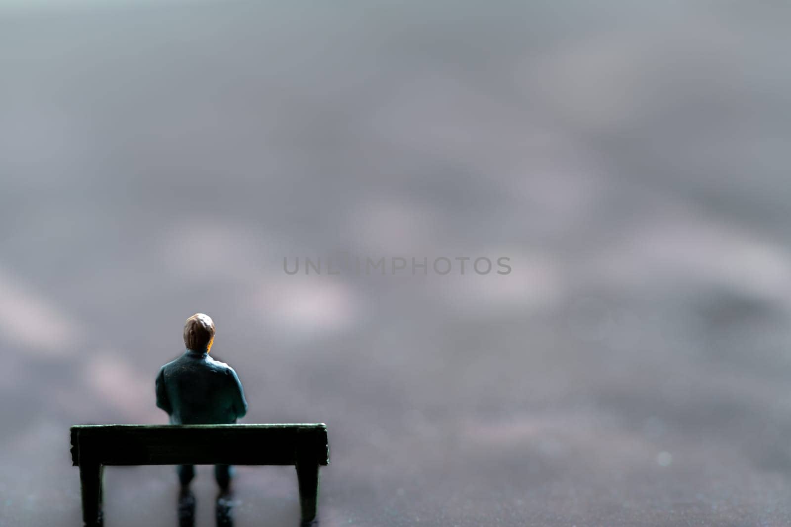 Miniature figurine posed as man sitting alone on a bench in surreal scenery, minimalist abstract concept image