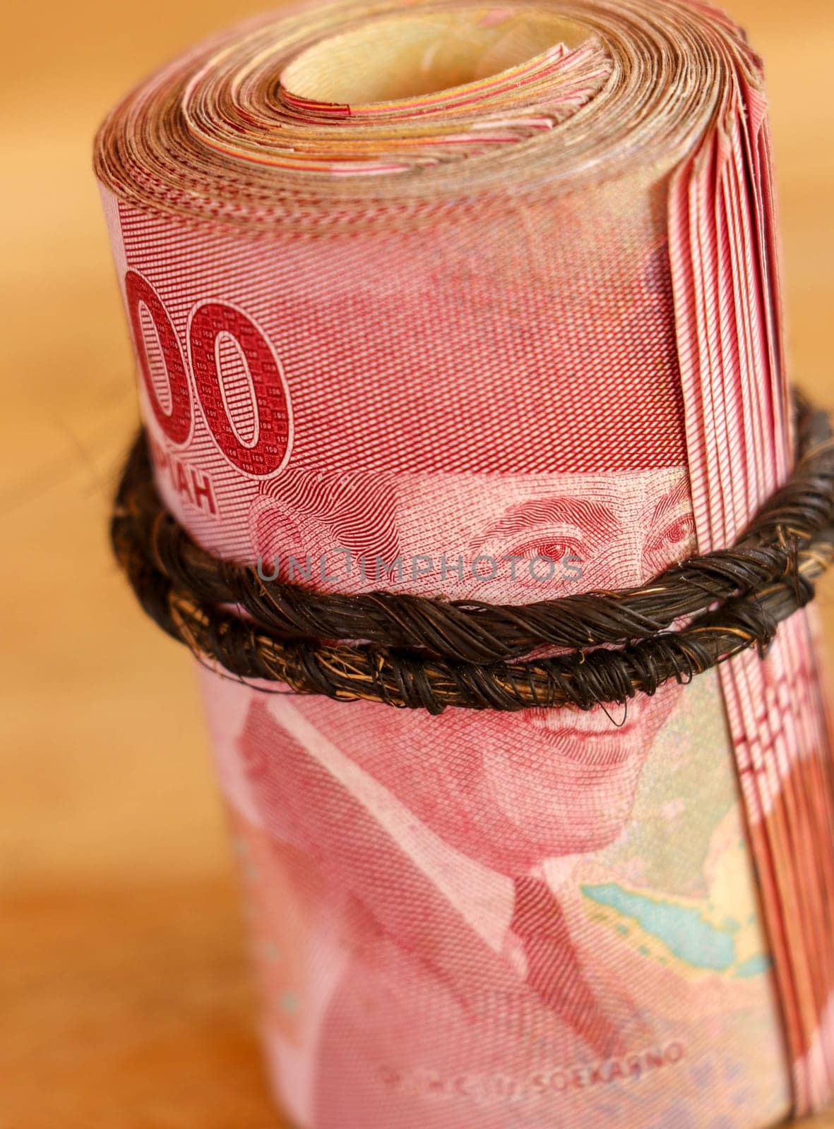 Indonesian banknotes rolled into a tube. IDR banknotes with a face value of 100000 rolled up with a brown string on an wooden background. Scrolled stack of dollar banknotes. Closeup, selective focus.