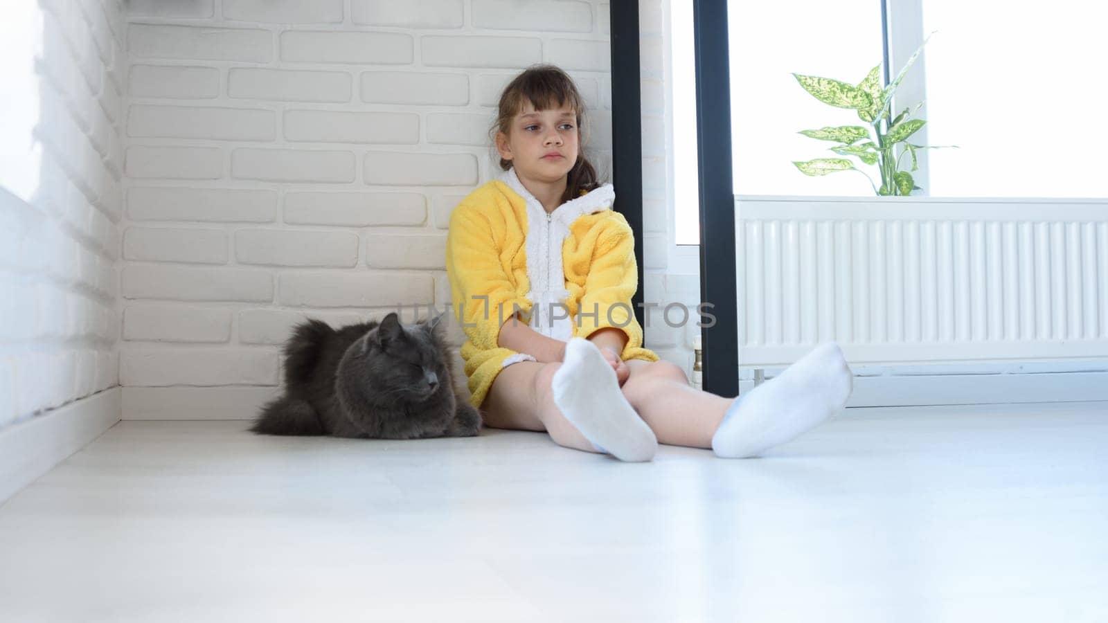 An upset girl in a yellow bathrobe sits in the corner of the room, a dark gray domestic cat sits nearby