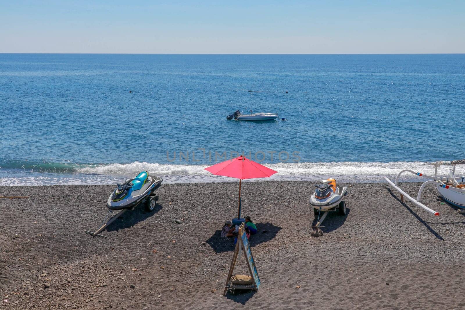 Jetski Seadoo parked on the beach. Black volcanic sand beach. In the background of the sea and moored boat. Red parasol.