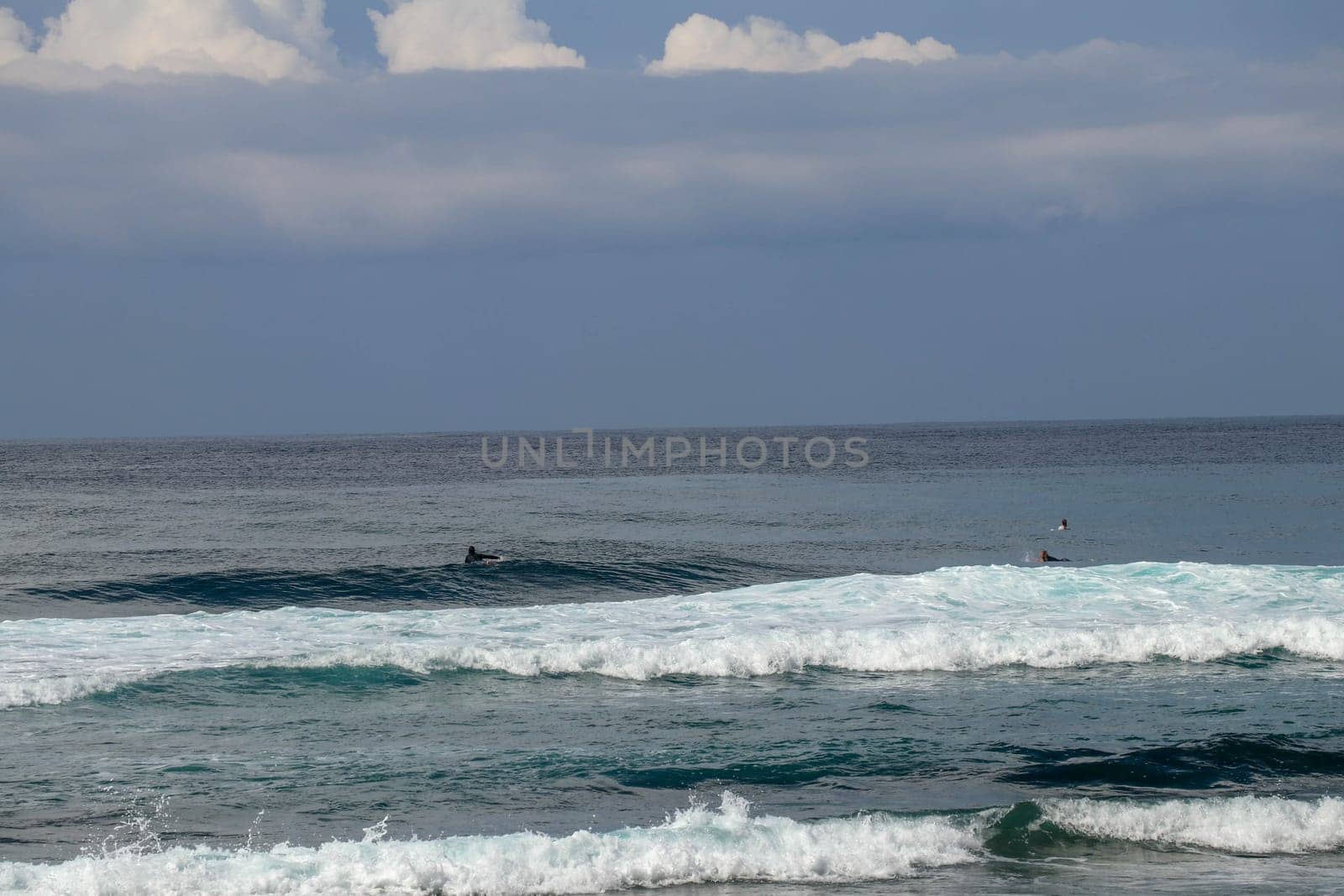 Surfers are waiting for a convenient wave on their surfboards. Tropical surfers paradise with Nyang Nyang beach on Bali island. Azure blue sky, sunny day. Wild waves are approaching the shore in rows.