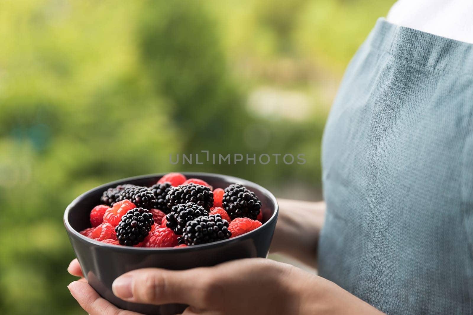 Female hands holding a bowl full of blackberries and raspberries. Unfocused background of trees.