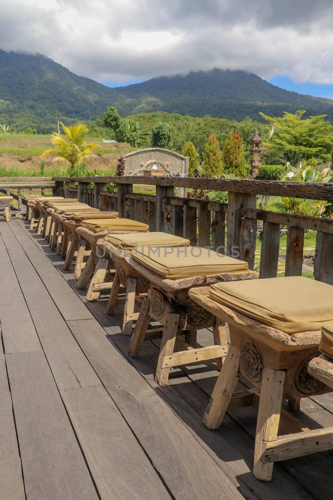 Row of wooden stools with cushions on terrace with floor made of planed wooden planks. It bumps from roughly worked beams. Background with mountains whose peaks are shrouded in thick clouds. Sunny day.
