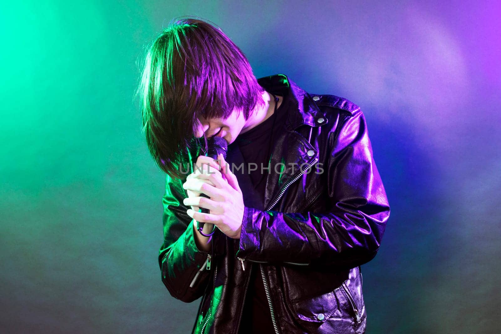 The young guy in leather jacket is singing rock music on illuminated stage on concert.