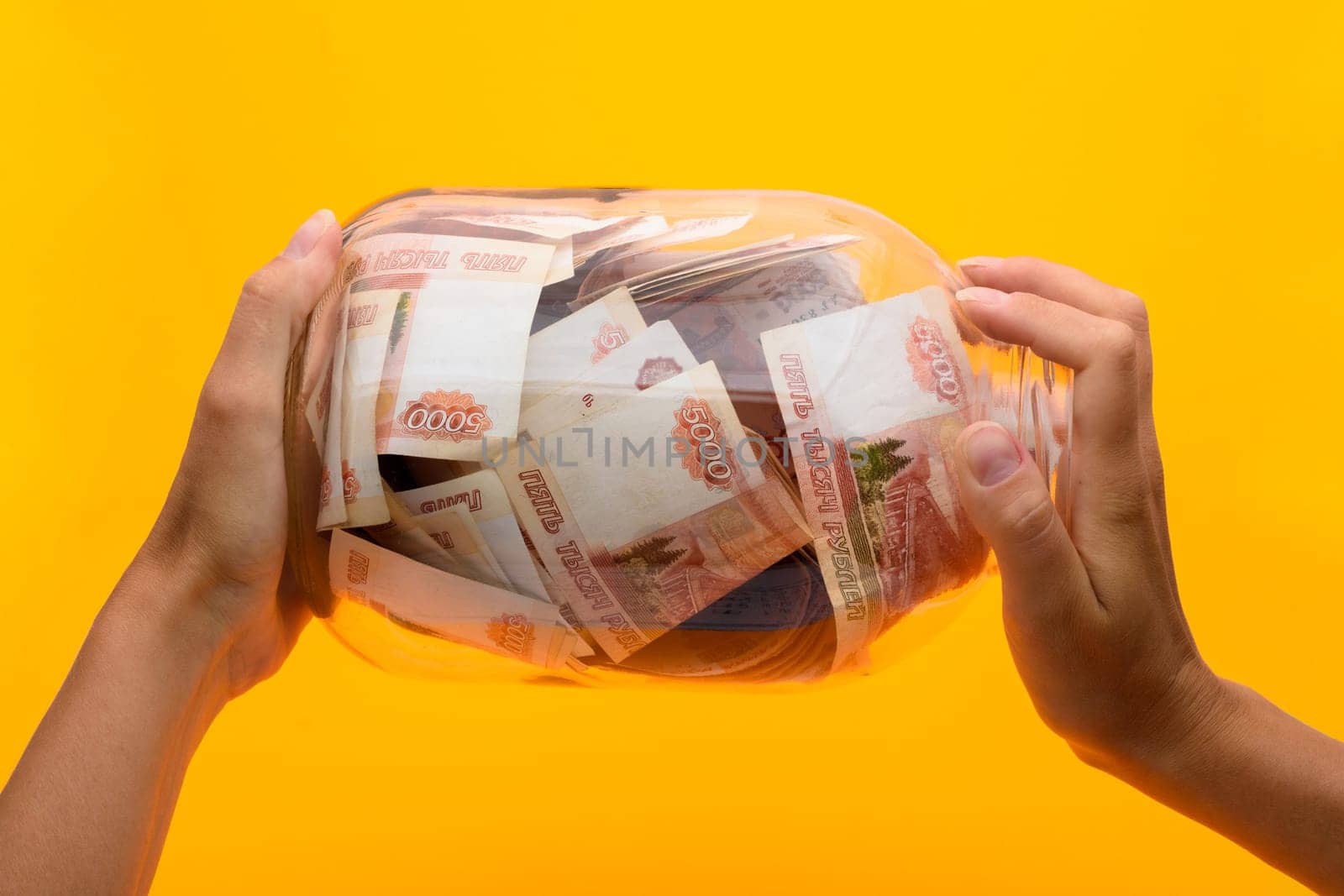 Hands holding a three-liter jar in which lie five thousandth bills, yellow background