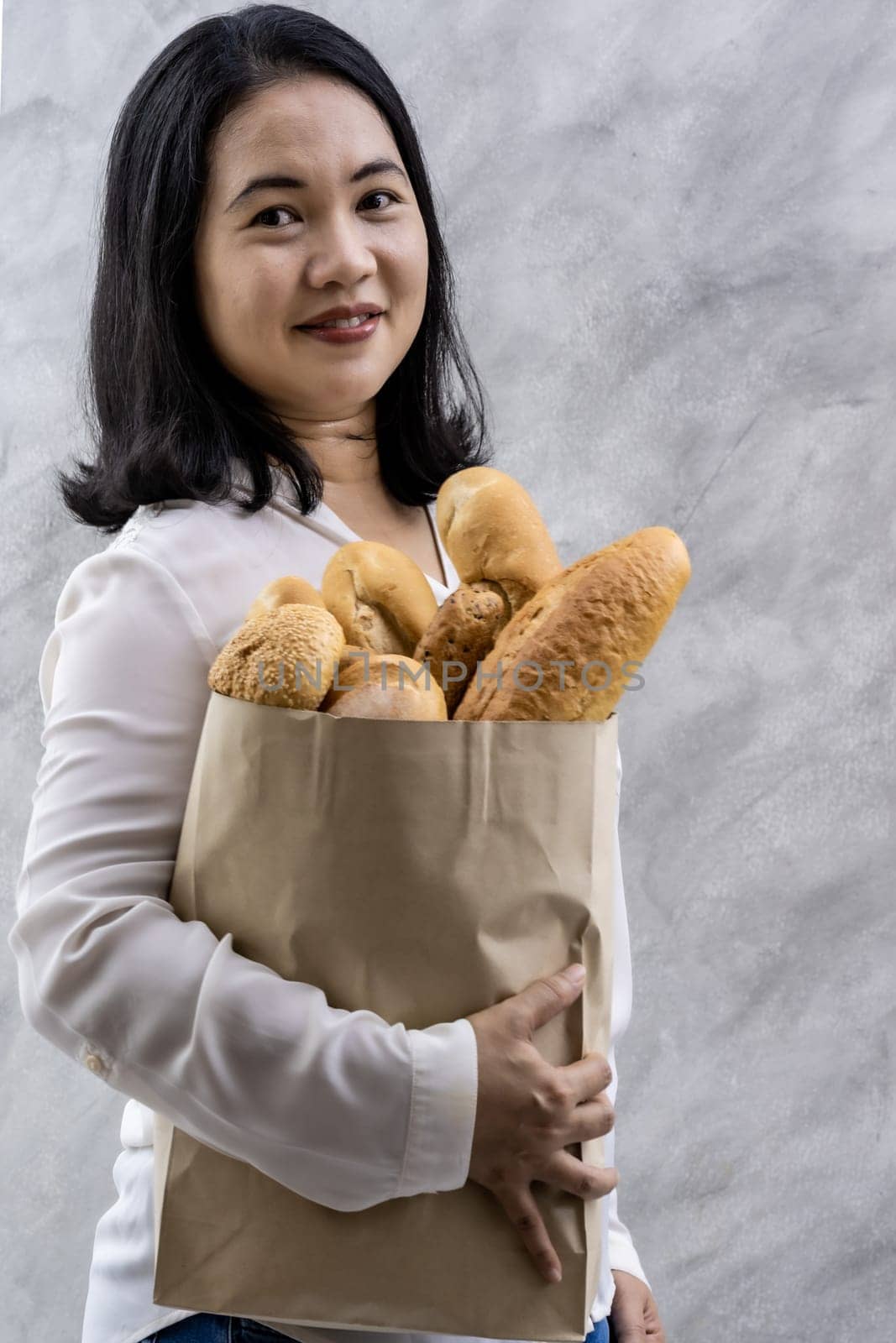 Portrait of asian smile woman housewife holding variety bread in disposable paper bag on grey vintage loft background. Bakery food and drink grocery and domestic life lifestyle concept for delivery.