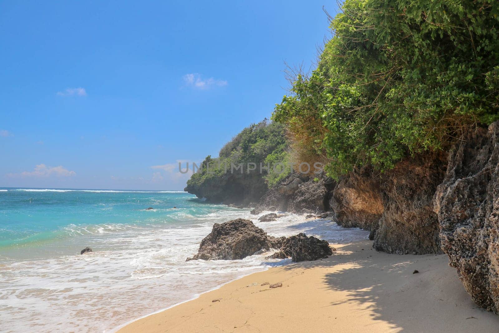 Pantai Gunung Payung beach with fine golden sand, Bali. Turquoise blue water turns into sea foam as the waves break against rocky cliffs of the coast. Sunny day with blue sky. Tourist natural concept.