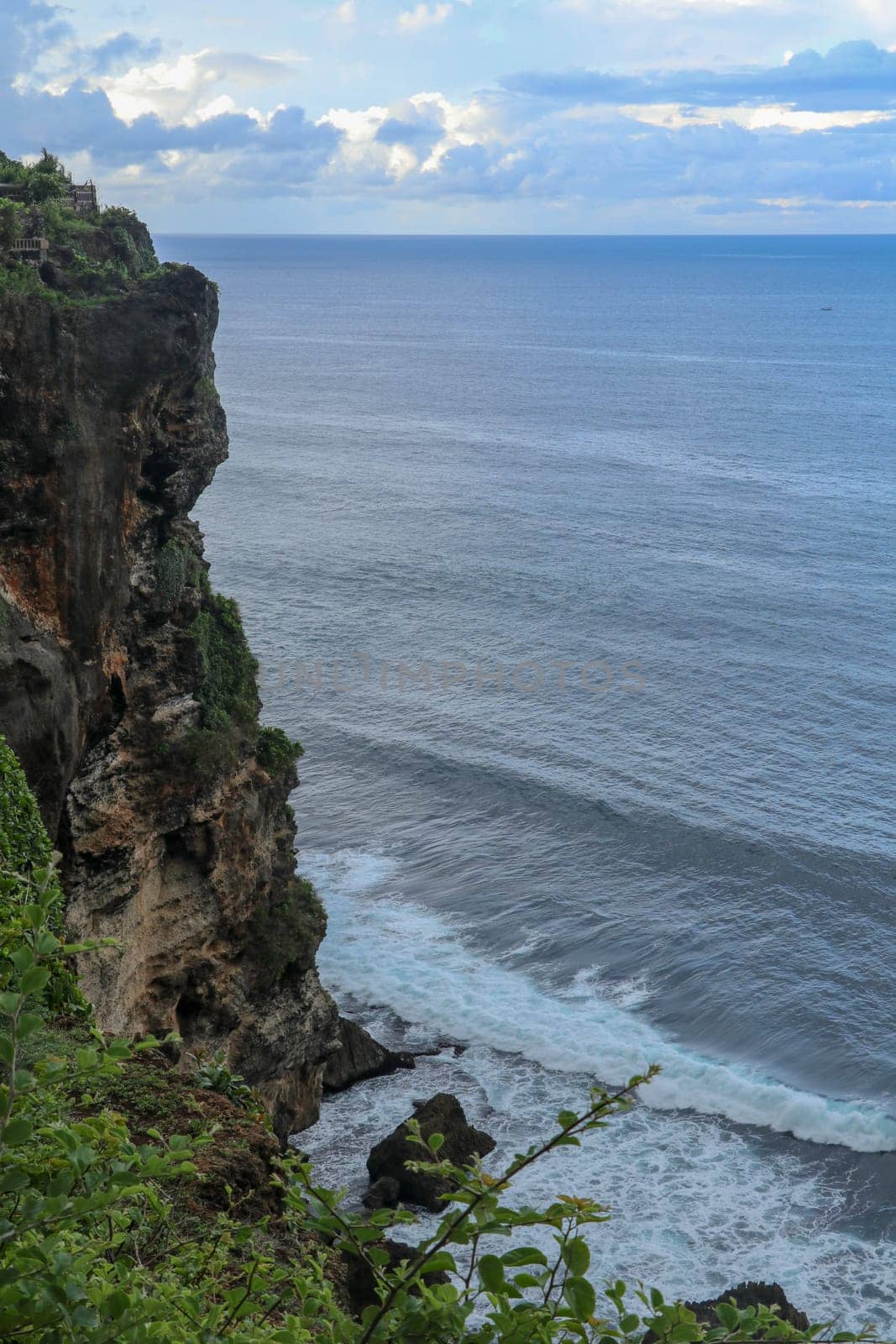 Overview panorama ocean shore, cliff. Sunset. Bali. Overwhelmed scene the flower and green-capped vertical cliff over the Indian ocean. South of Bali island, Pura Luhur Uluwatu Temple. Indonesia.