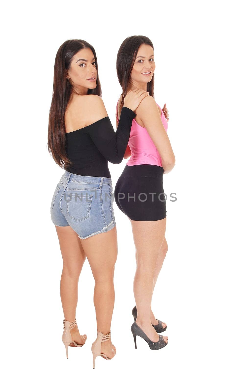 A two happy young slim women standing in the studio in shorts behind.each other, looking at camera, isolated for white background.
