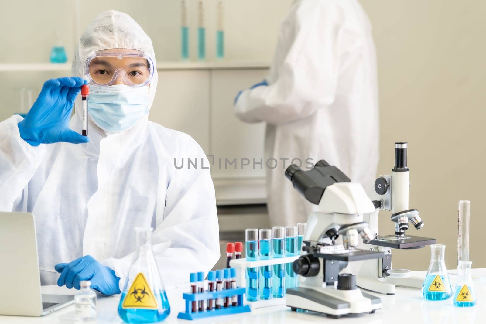 Scientists hold blood sample test tube and examine for his research and develop vaccine for coronavirus covid-19 pandemic with his colleague in background. Medical Science and technology concept.