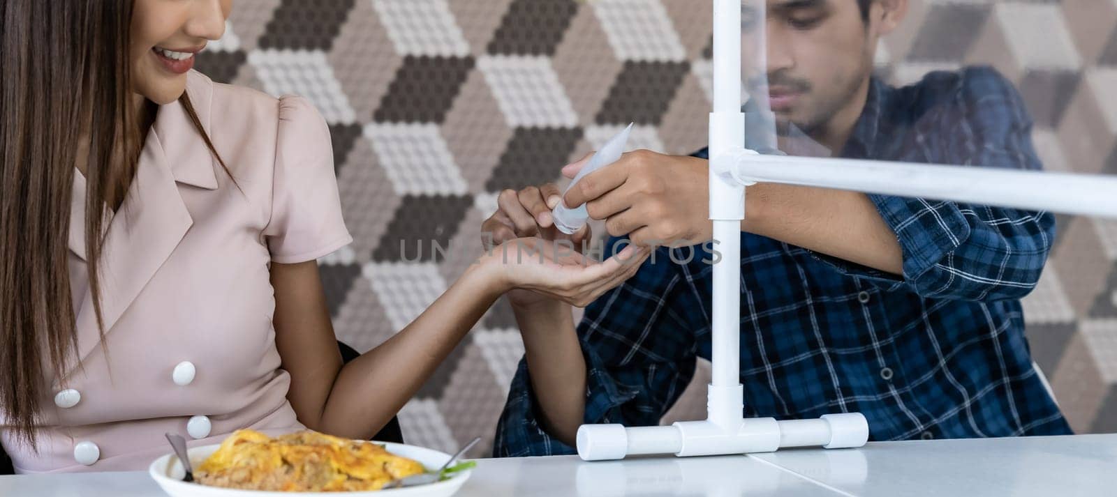 Panoramic asian man put alcohol gel hand sanitizer to his partner before eating at new normal social distance restaurant with table partition. Restaurant new normal lifestyle. Web banner crop.