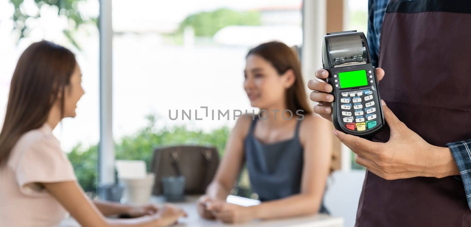 Panoramic close up of waiter hand hold credit card reader for contactless payment with customers in restaurant background. New normal restaurant contactless payment concept.
