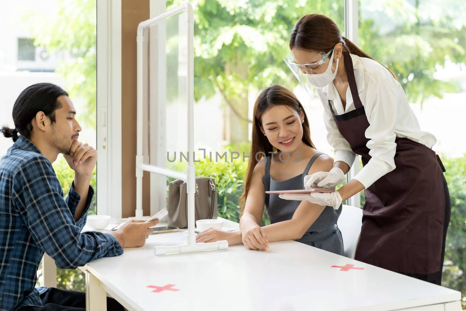 Asian waitress wear facemask and face shield using tablet to show restaurant electronic menu to reduce touching and recommend to customer. New normal technology and lifestyle food and dining concept.