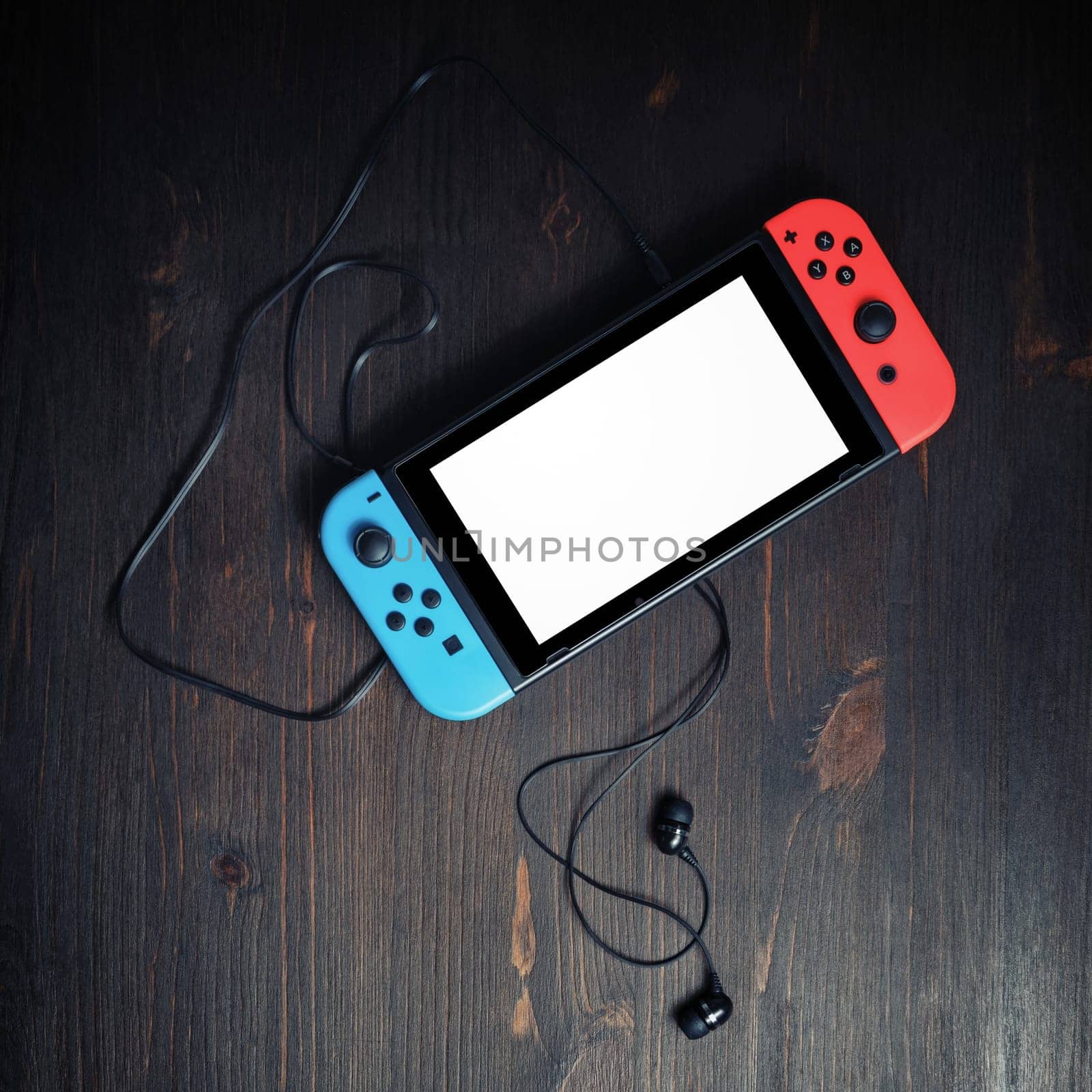 Minsk, Belarus - May 07, 2020: Nintendo Switch game console with blank white screen and headphones on vintage wooden background. Flat lay.