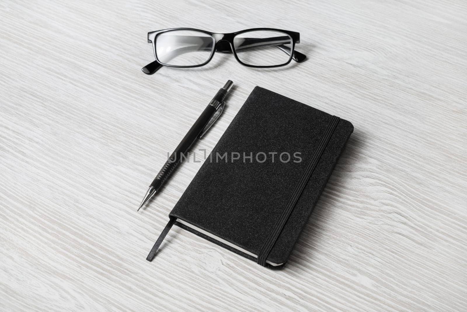 Photo of closed blank black notepad, glasses and mechanical pencil on light wood table background. Template for placing your design.