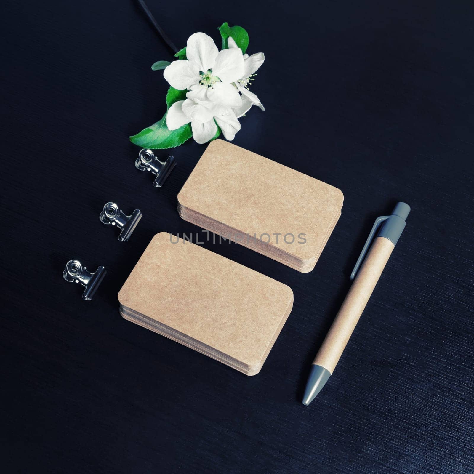 Blank kraft paper business cards, pen and flowers on black wooden background. ID template. Mock-up for branding identity for designers.