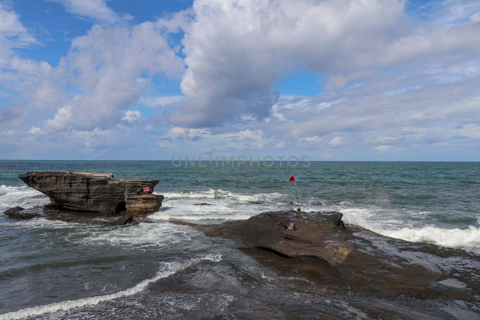 Sea surf and waves hit the rock cliffs at Tanah Lot Temple. Sea water turns into white foam. Red warning flag highlights dangers of ocean. Unbridled ocean energy. Natural background for your project.