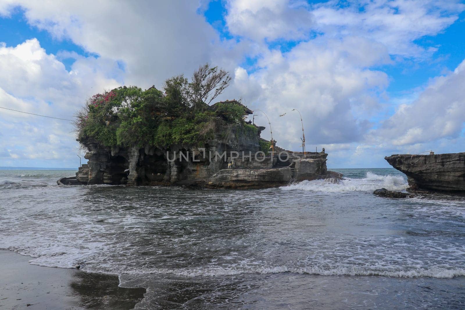Waves shatter on a cliff at the top of which is the Hindu temple of Tanah Lot. Temple built on a rock in the sea off the coast of Bali island, Indonesia. Lush tropical vegetation on top of a cliff.