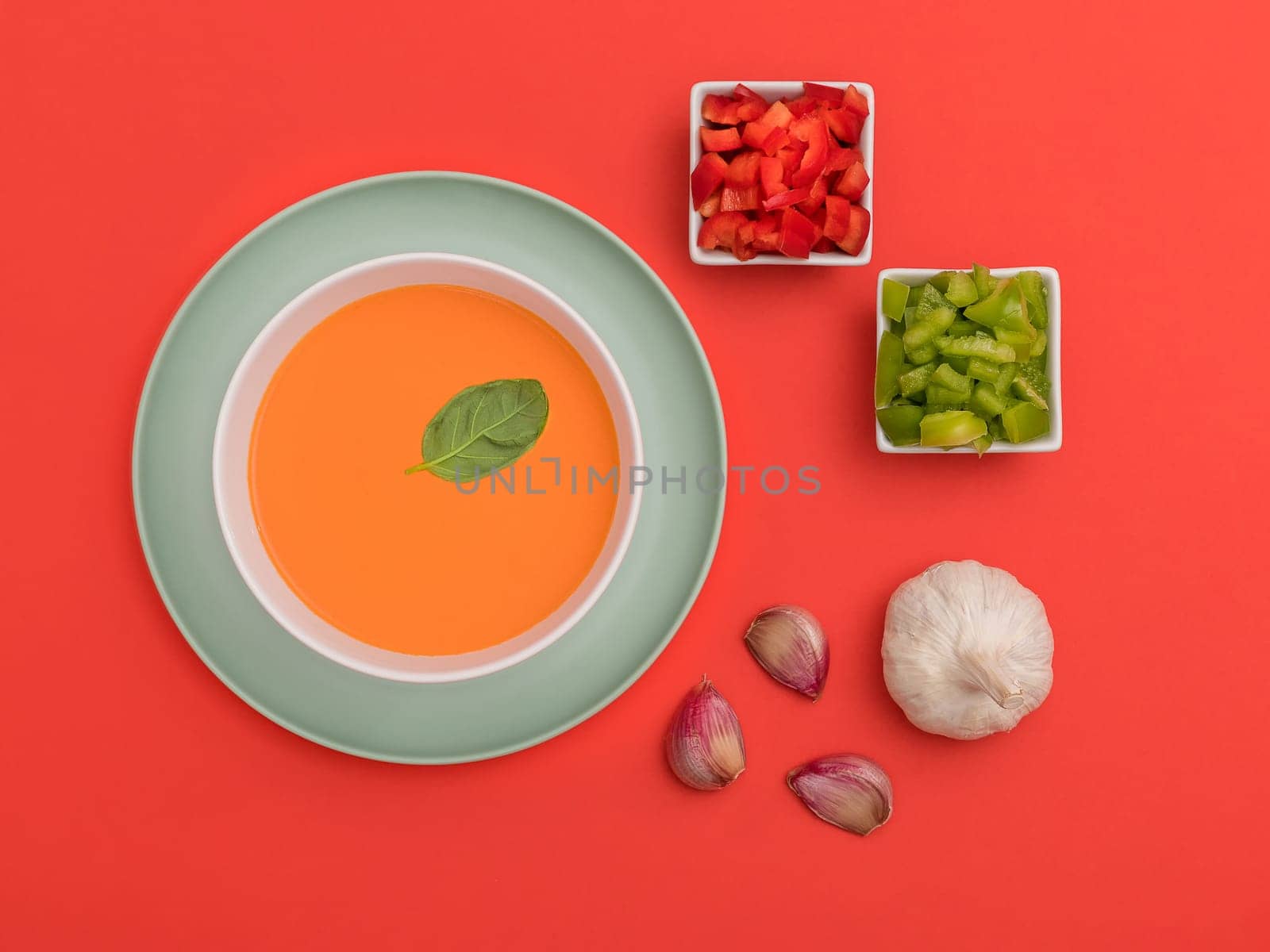 Gazpacho white bowl on a blue plate with garlic cloves and red and green peppers on red background. Top view. Copy space.