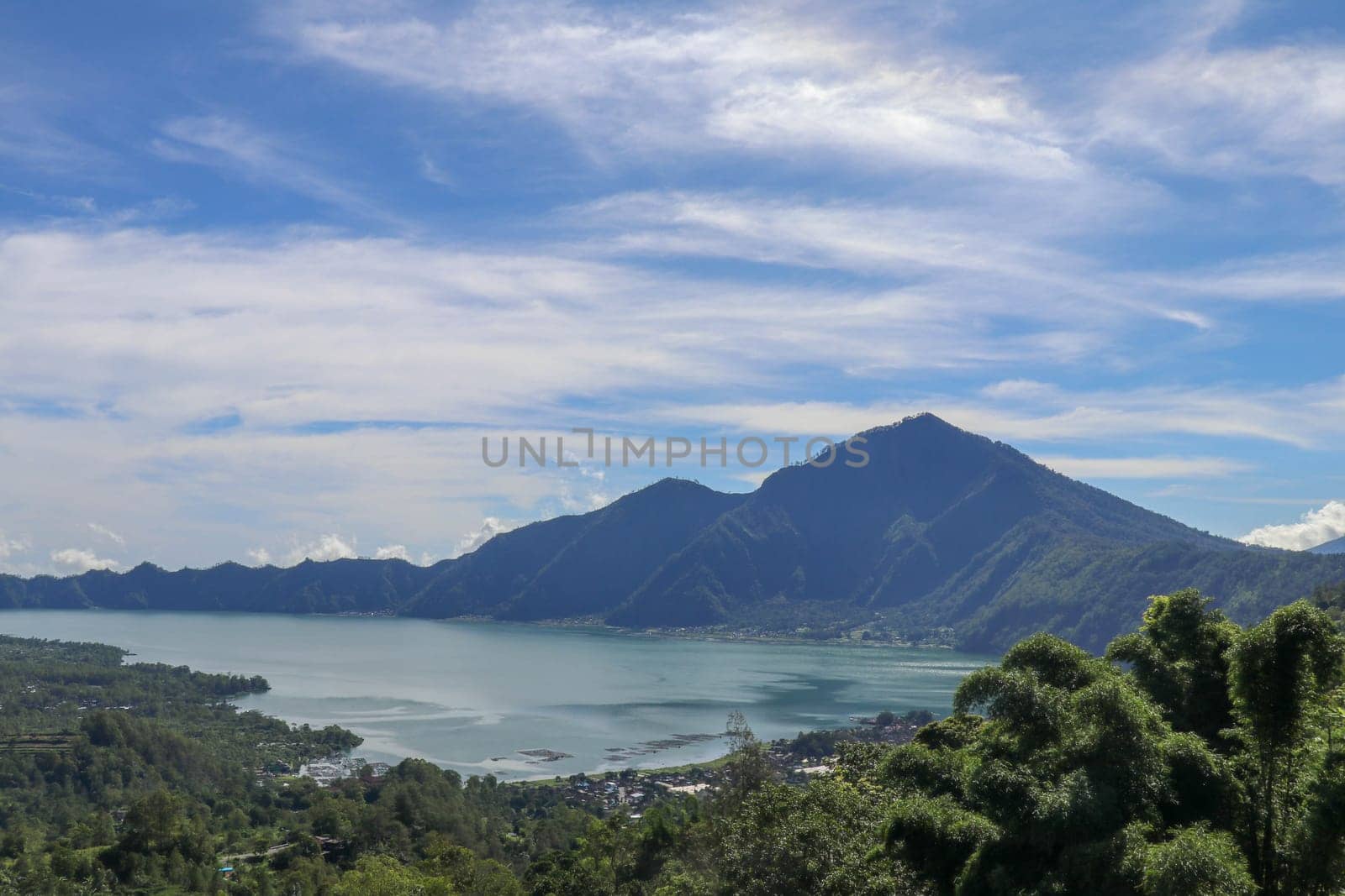 Mountain lake in the volcanic caldera of Batur under Mount Abang on the island of Bali, Indonesia. Quiet level of a large drinking water reservoir. High mountain peaks rise above the lake. Blue sky.