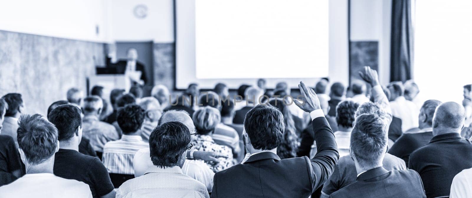 I have a question. Group of business people sitting at the chairs in conference hall. Businessman raising his arm. Conference and Presentation. Business and Entrepreneurship. Blue toned.