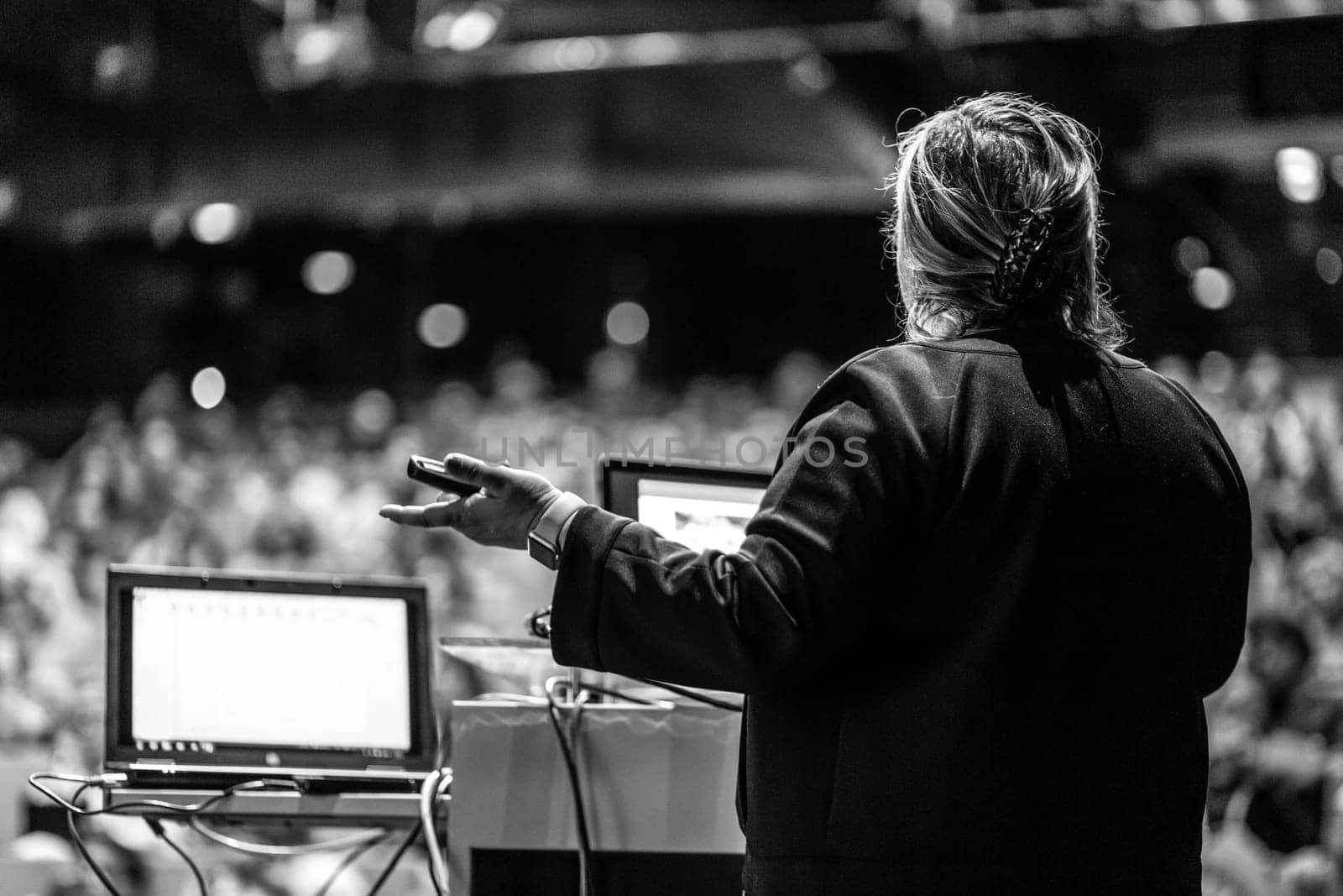 Female speaker giving a talk on corporate business conference. Unrecognizable people in audience at conference hall. Business and Entrepreneurship event. Black and white image.