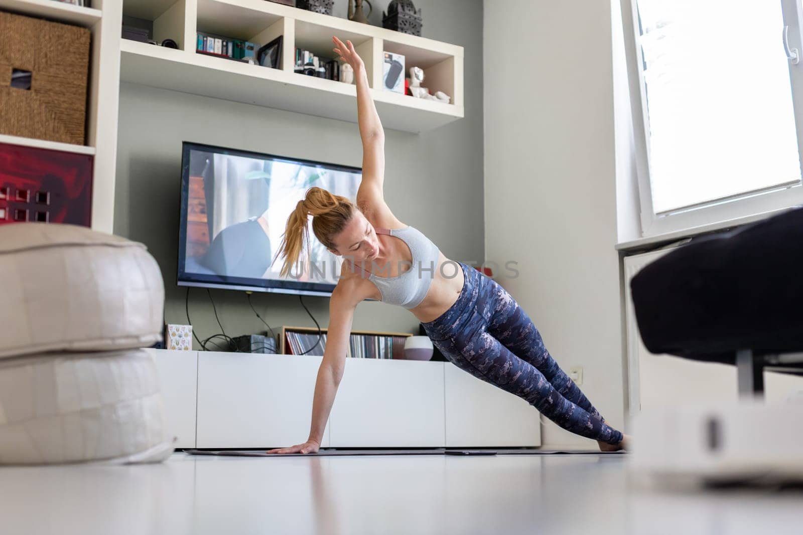 Attractive sporty woman working out at home, doing pilates exercise in front of television in her living room. Social distancing. Stay healthy and stay at home during corona virus pandemic.