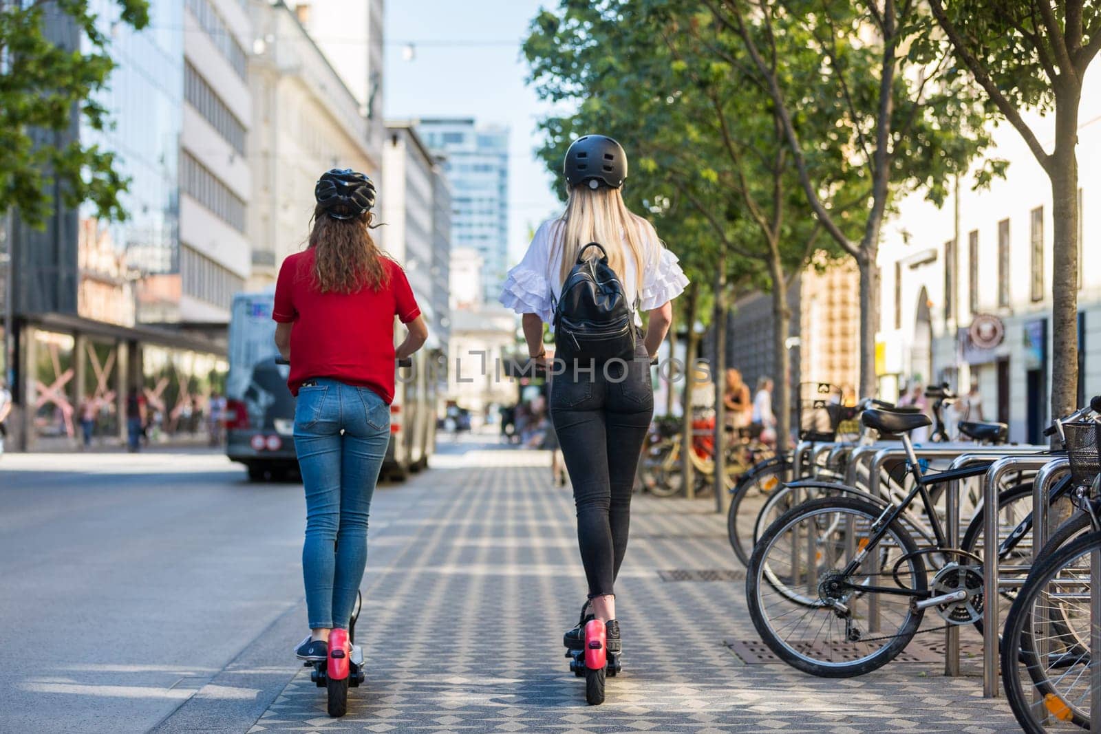 Rear view of trendy fashinable teenager girls riding public rental electric scooters in urban city environment. New eco-friendly modern public city transport in Ljubljana, Slovenia.
