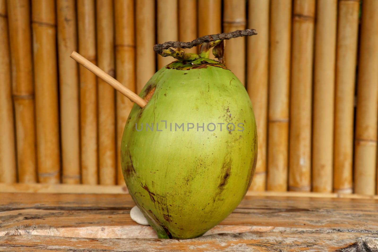 Fresh young coconut with bamboo straw on table with massive wooden board. Healthy natural refreshment full of energy and vitamins. In the background wall of bamboo trunks. Nice tropical concept.