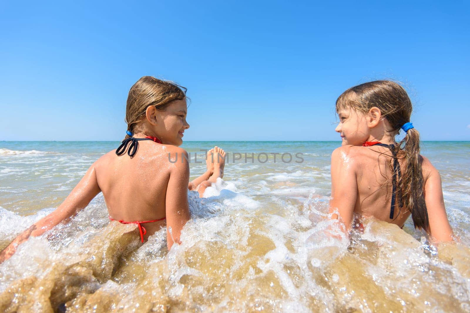 Children sit in water on a sandy beach and look at each other happily