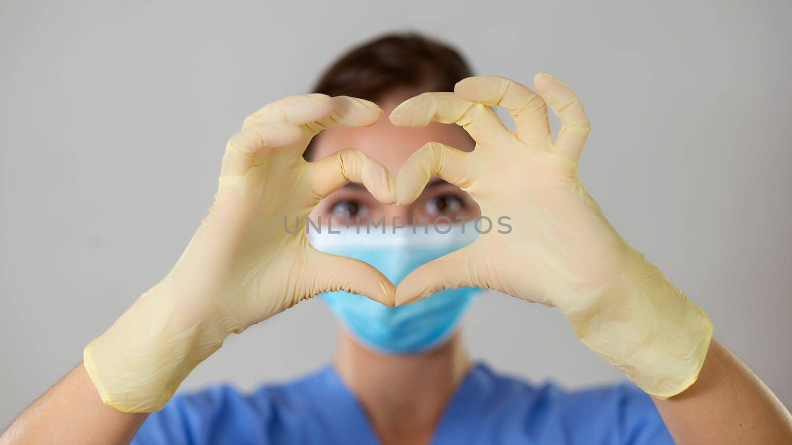 Young girl in a blue medical attire looking at the camera through a heart shape formed with gloved fingers. Picture of a close-up heart symbol.