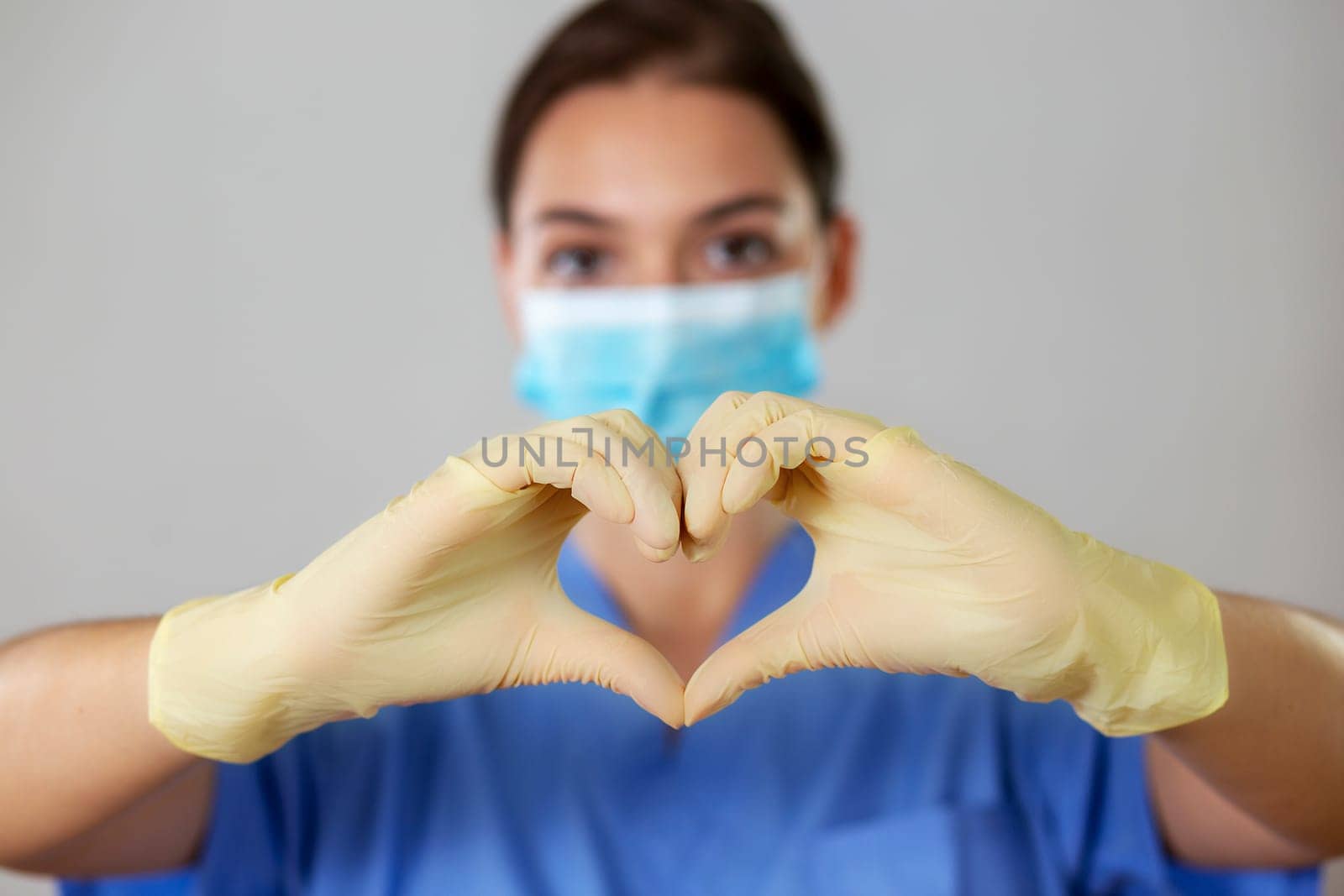 Young girl in a medical mask showing the symbol of a heart with gloved hands in front of her on a light studio background. Front view of caucasian woman in medical uniform.
