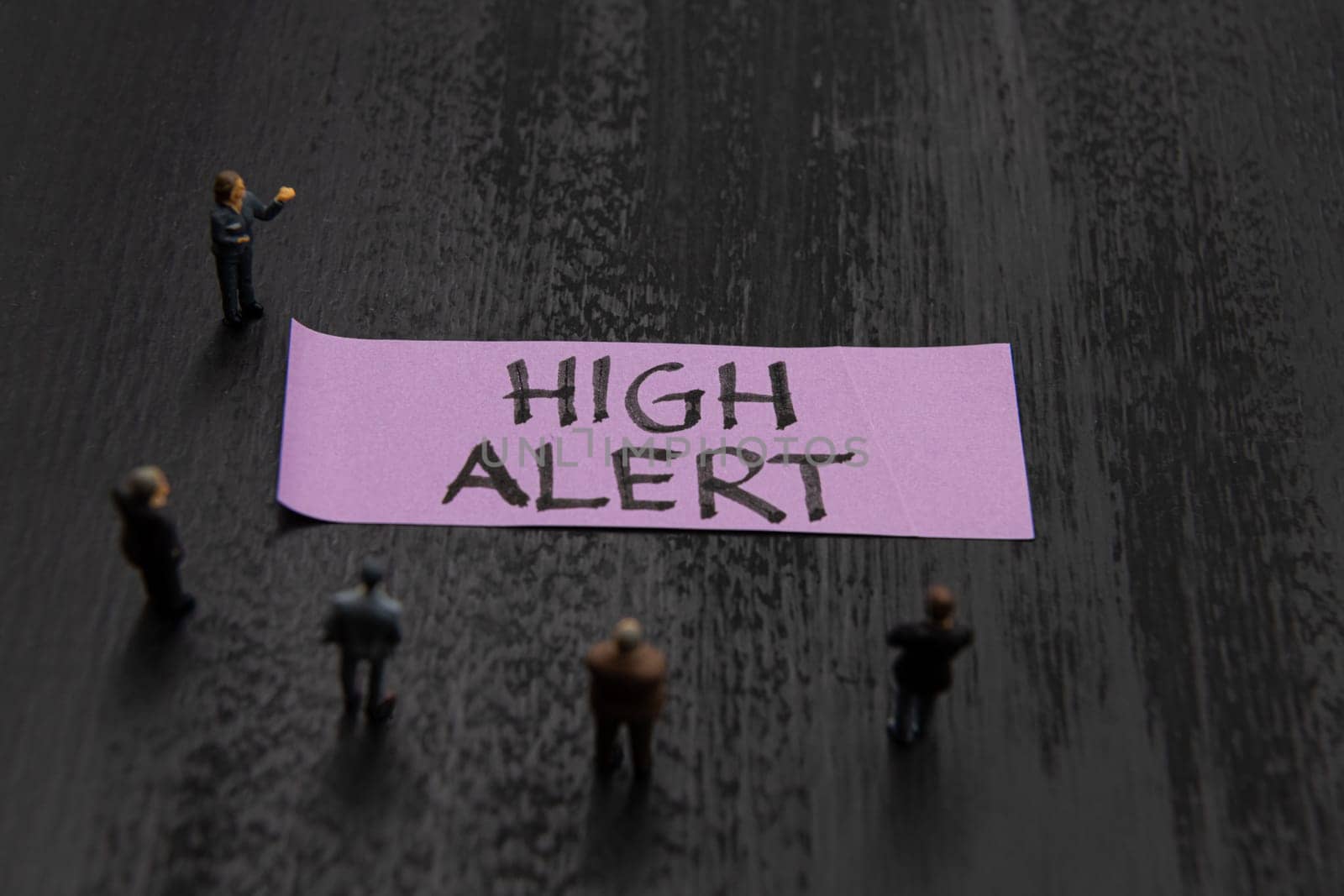 Strategy meeting with miniature figurines posed as business people standing around post-it note with High Alert message, minimalist abstract concept image with focus on text