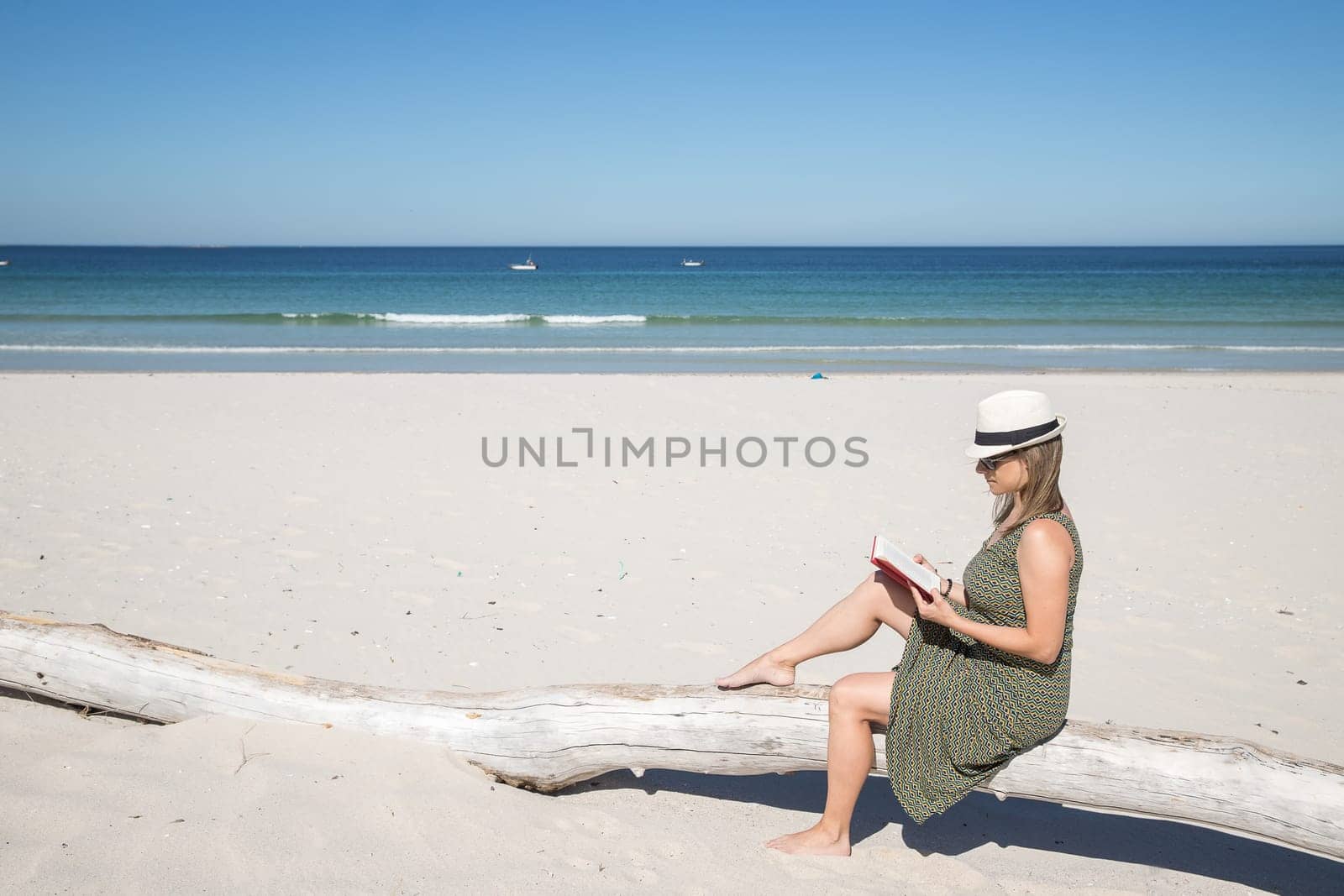 Middle-aged blonde woman sitting on a tree trunk n the beach reading a book with the sea on the background