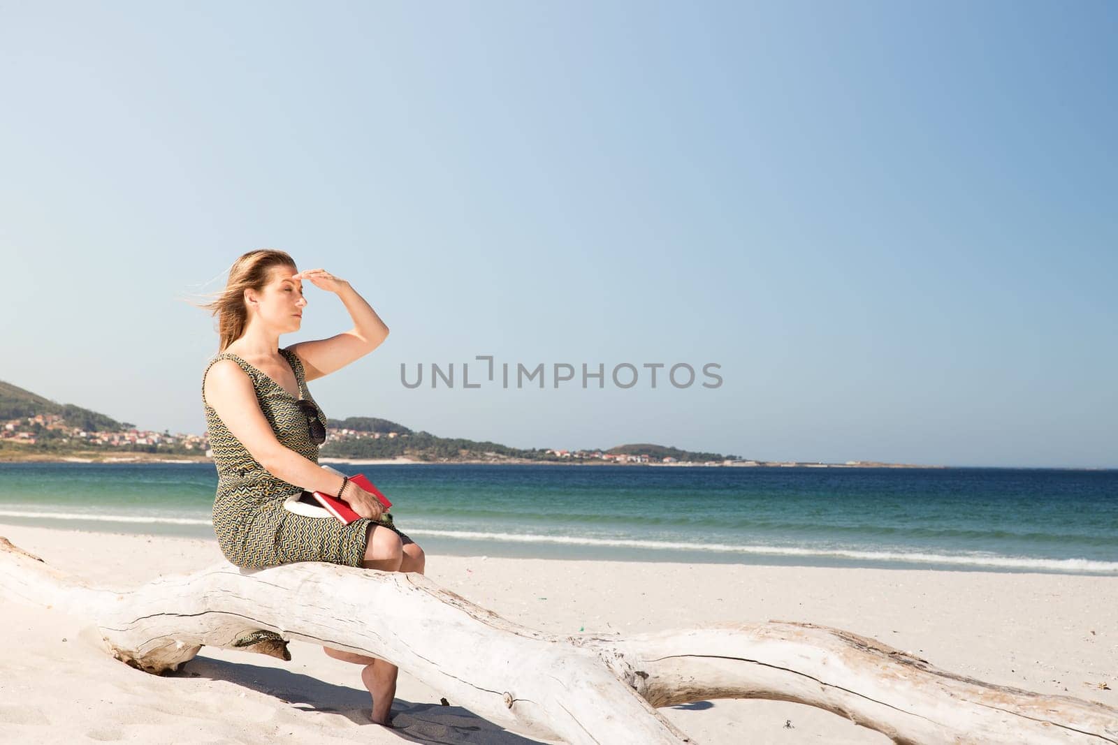 A middle-aged blonde woman sitting on a tree trunk at the beach, protecting herself from the sun with her hand to look in the distance and with a book on her legs