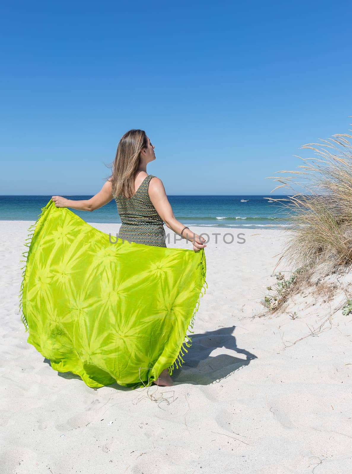 A middle-aged blonde woman on her back holding a green sarong on the beach