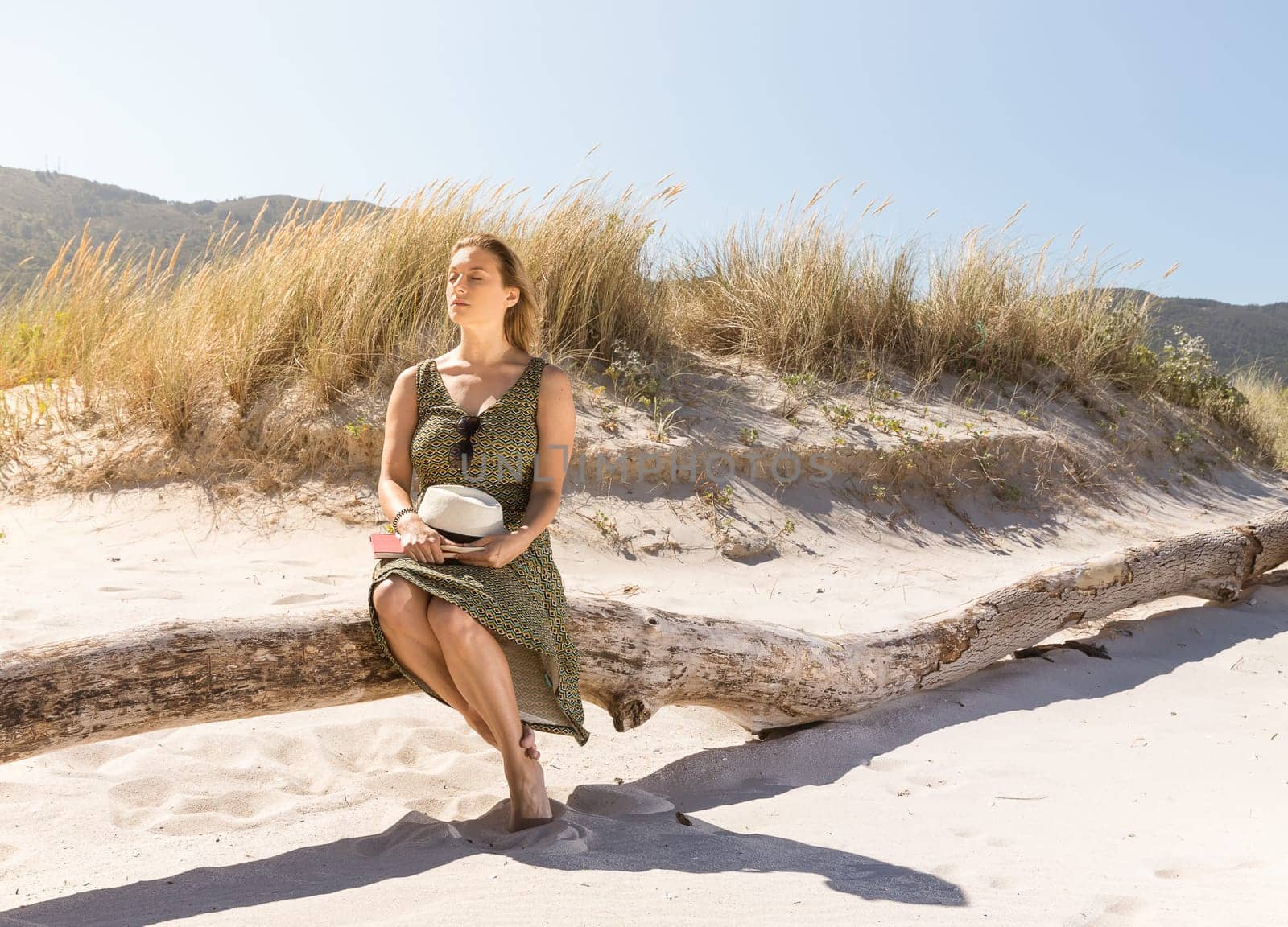 A middle-aged blonde woman sitting thoughtfully on a tree trunk at the beach with a hat and a book on her legs