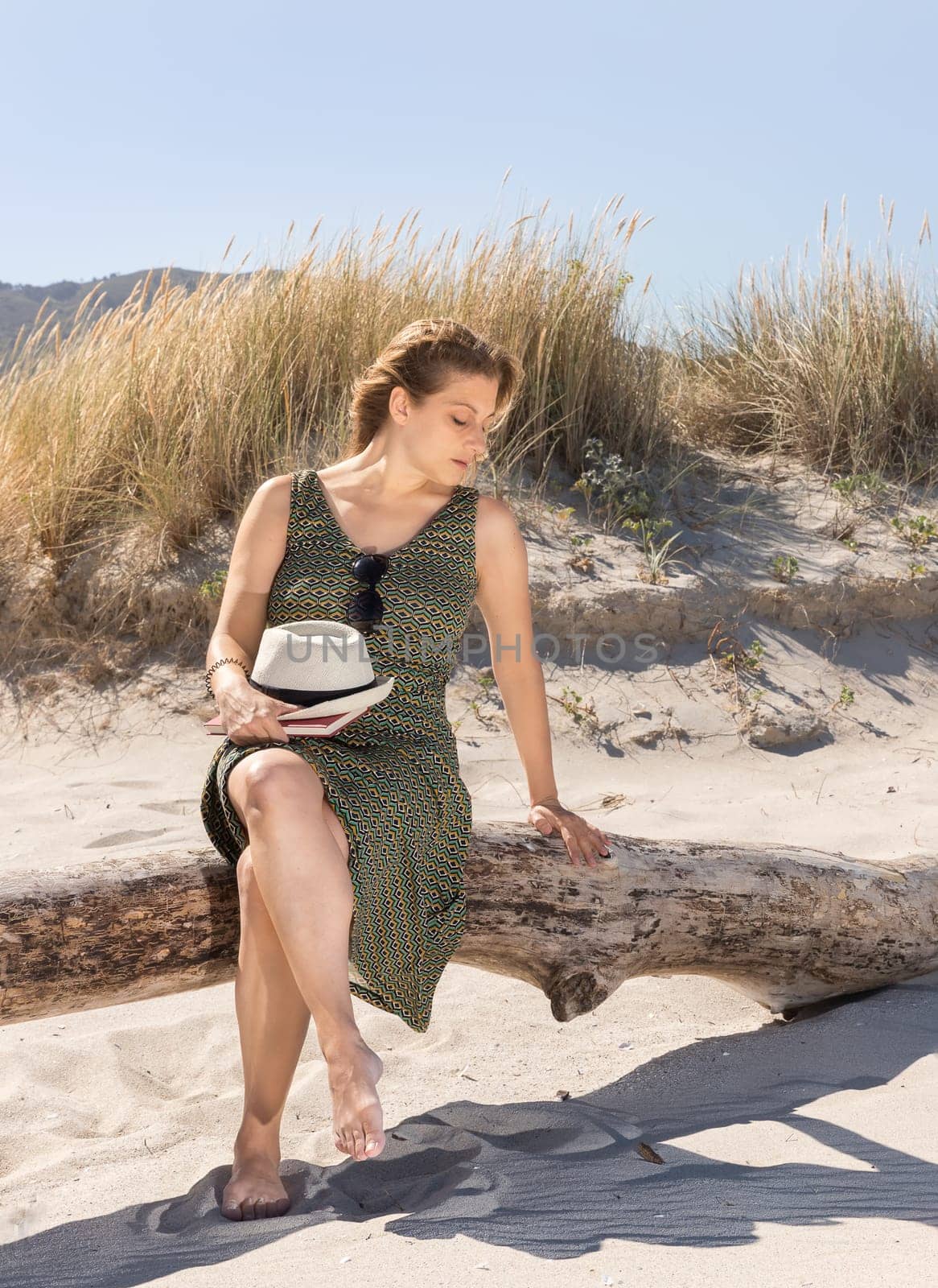 A middle-aged blonde woman sitting thoughtfully on a tree trunk at the beach with a hat and a book on her legs