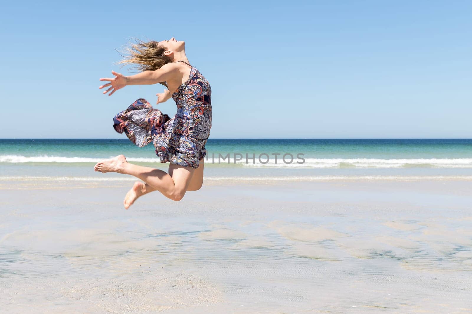 Middle-aged blonde woman in a green dress jumping with her arms open on the beach