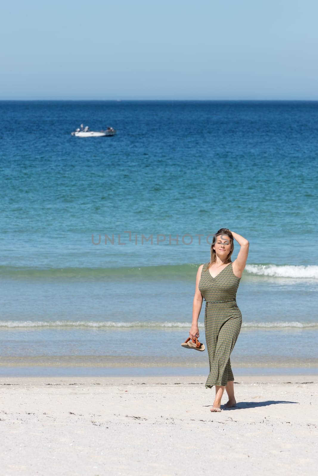 Middle-aged blonde woman in a green dress walking barefoot on the beach on the beach with her sandals in one hand