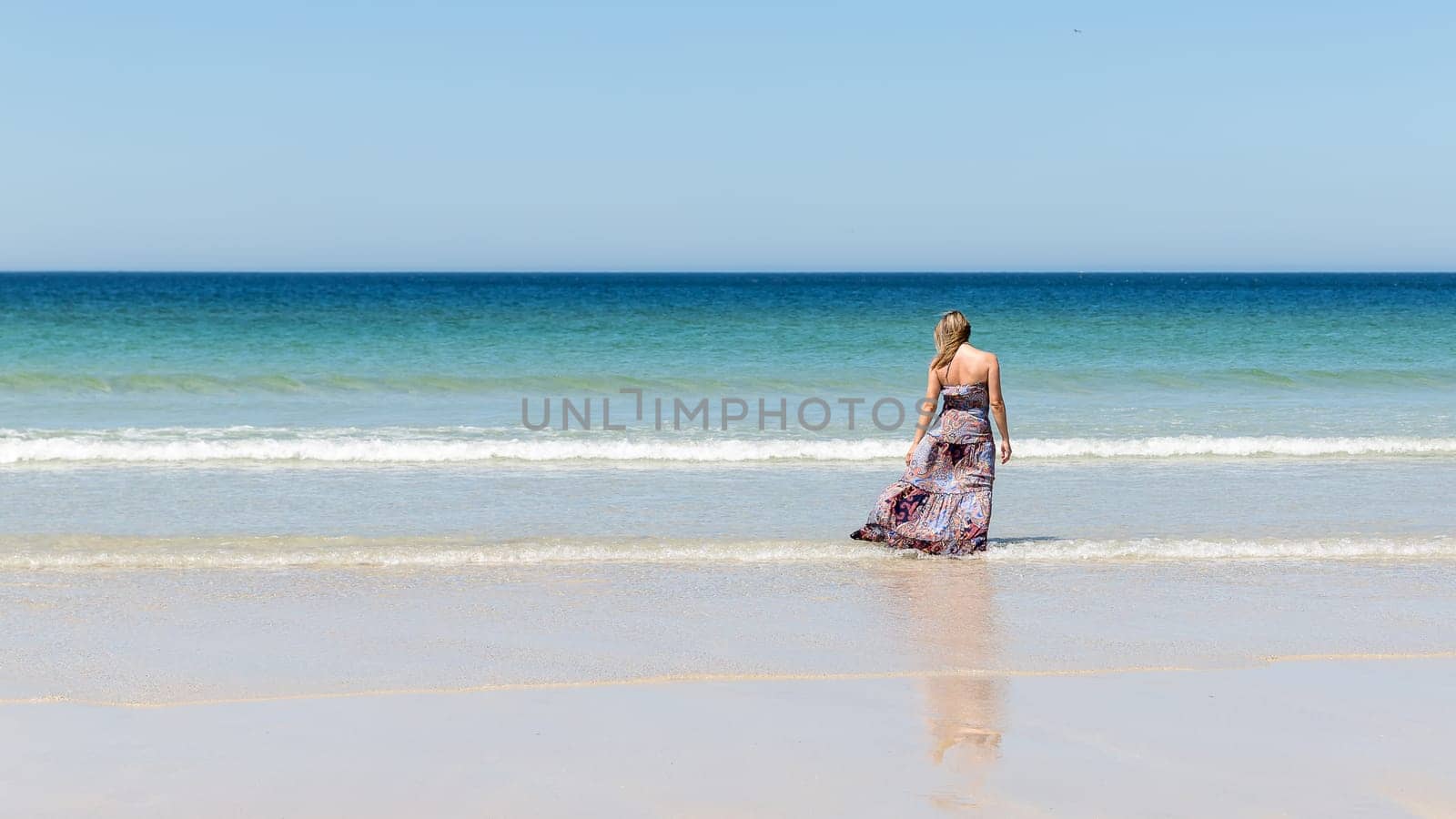 Middle-aged blonde woman in a long dress relaxes on the beach facing the sea