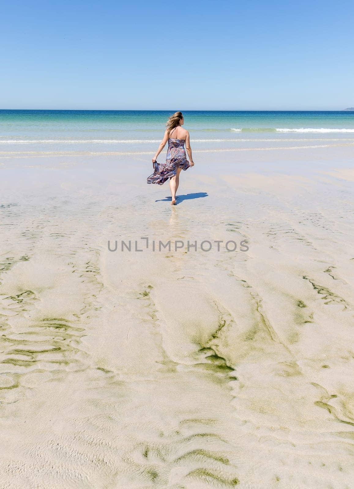 Middle-aged blonde woman in a long dress walking on the beach towards the sea